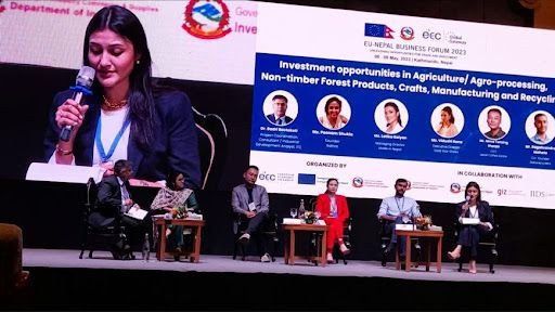 Latika Golyan presenting at the EU-Nepal Business Forum 2022, with a panel of individuals seated on stage behind her, in a conference hall setting with a large screen displaying information about investment opportunities.